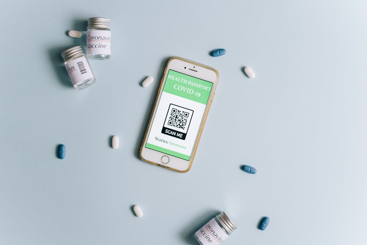 Image of a smartphone displaying a health passport alongside COVID-19 vaccine vials and pills on a light blue background.